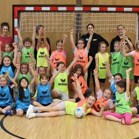 Eine Gruppe von Kindern in Sporttrikots mit Erima-Logos posiert für ein Foto in einer Turnhalle. Sie sitzen und stehen vor einem rot-weißen Tor.
