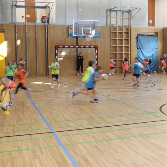 Kinder spielen Fußball in einer Halle. Einige rennen mit Bällen, während andere in der Nähe des Tors sind. Ein Trainer steht in der Nähe.