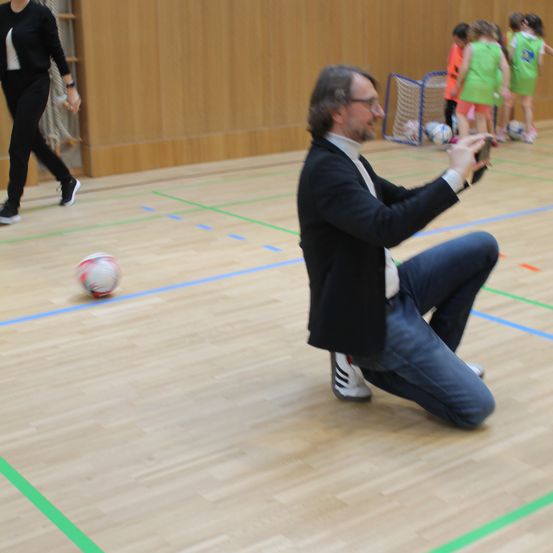 Ein Mann kniet auf einem Spielfeld, möglicherweise als Trainer einer Fußballpartie. In der Nähe geht eine Frau und hält einen Ball. Im Hintergrund spielen Kinder Fußball in der Nähe eines Tores.