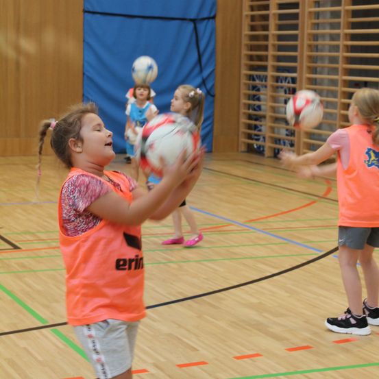 Drinnen spielen junge Mädchen mit Fußballen, eine hält einen Ball, während eine andere versucht, ihn zu fangen. Im Hintergrund sind blaue Wände und Holzregale zu sehen.