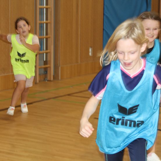 Drei junge Mädchen spielen Basketball in einer Turnhalle. Zwei von ihnen tragen blaue Trikots mit dem Wort 'erima' darauf.