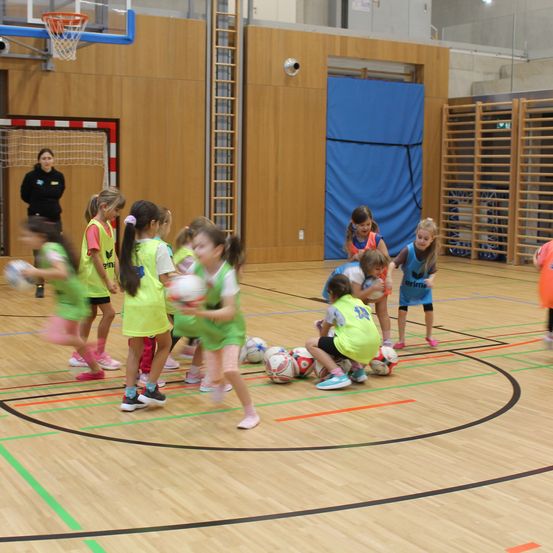 Eine Gruppe junger Mädchen in grünen Trikots spielt Hallenfußball. Sie stehen und knien auf dem Spielfeld und halten Fußballbälle. Ein Basketballkorb und Netz sind an der Wand montiert. Eine Frau steht in der Nähe der Wand.