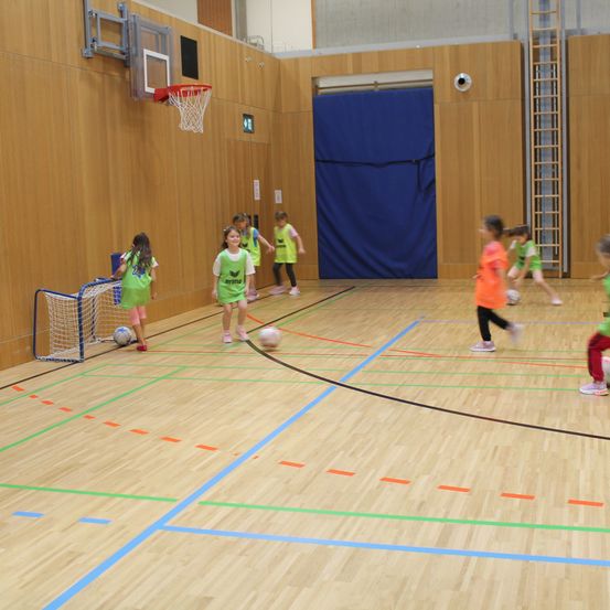 Mehrere Kinder in grünen Trikots spielen drinnen Fußball, einige mit Bällen. Ein Basketballkorb ist an der Wand montiert. Der Boden hat farbige Markierungen.