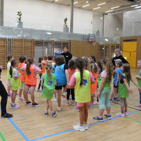 In einer Turnhalle sind mehrere junge Mädchen in einem Kreis versammelt. Sie tragen farbenfrohe Westen und Turnschuhe. Eine erwachsene Frau steht vor ihnen und gibt wahrscheinlich Anweisungen. Im Hintergrund befinden sich Holzwände und eine gelbe Tür.