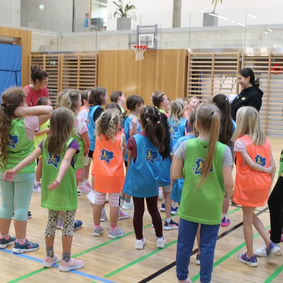 Eine Gruppe junger Mädchen in passenden Westchen versammelt sich auf einem Basketballplatz und hört einer Frau in einer schwarzen Jacke zu. Der Platz hat einen Basketballkorb und Holzwände.