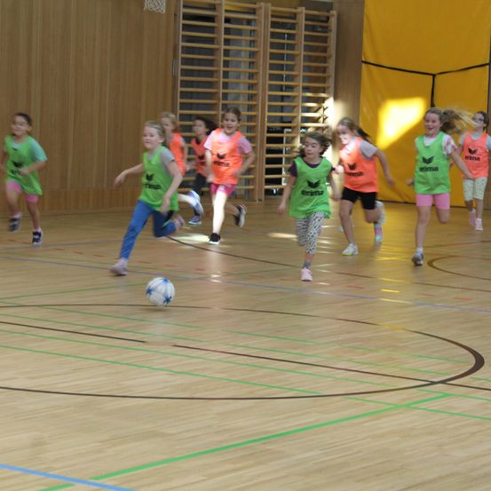 Eine Gruppe junger Mädchen in grünen und orangefarbenen Trikots spielt Fußball in einer Halle. Das Mädchen in Grün rennt auf den Ball zu, während andere ihm folgen. Der Spielfeldboden hat blaue und grüne Markierungen.