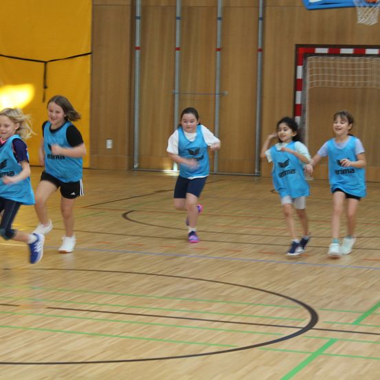 Fünf junge Mädchen in passenden blauen Trikots laufen lächelnd und engagiert auf einem Basketballplatz, während sie ein Spiel spielen.