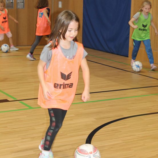 Eine Gruppe junger Mädchen in orangefarbenen Westen spielt Fußball auf einem Hallenplatz. Das Mädchen in der Mitte trägt eine orangefarbene Weste mit dem Wort 'erima' darauf.
