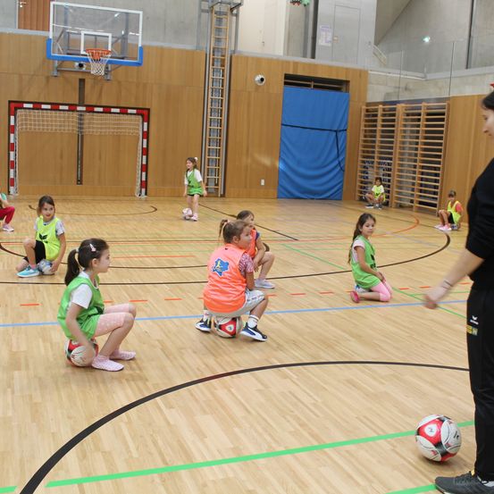 Eine Gruppe junger Mädchen trainiert auf einem Hallenfußballplatz mit einer Lehrerin. Einige sitzen mit Fußballbällen, während andere in einer Reihe stehen. Der Platz hat Holzböden und Markierungen.