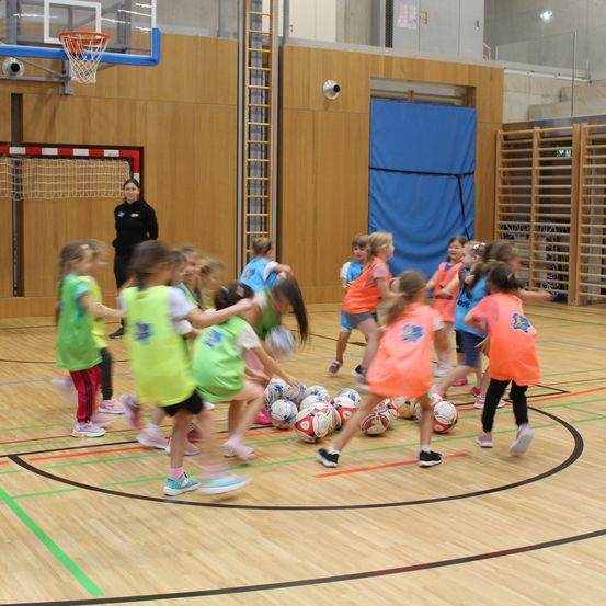 Eine Gruppe von Kindern in Sportbekleidung spielt auf einem Hallenplatz mit Fußballbällen, beaufsichtigt von einem Erwachsenen.