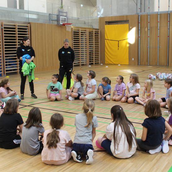 Eine Gruppe von Mädchen sitzt in einem Kreis auf einem Basketballplatz, während zwei Frauen in der Nähe stehen, eine hält ein Shirt. Sie scheinen an einem Gespräch oder einer Anweisung teilzunehmen.