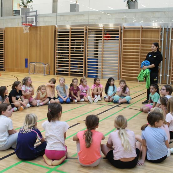 Eine Gruppe junger Mädchen sitzt in einem Kreis in einer Turnhalle und hört einer Frau zu, die steht und ein blaues Tuch hält. Die Turnhalle hat Holzböden, einen Basketballkorb und Holzregale.