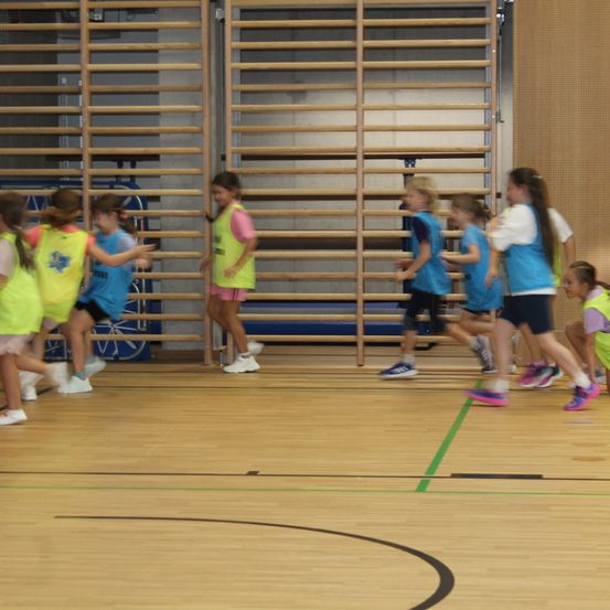 Mehrere Mädchen laufen auf einem Spielfeld und tragen gelbe und blaue Trikots. Hinter ihnen sind Holzstangen an der Wand montiert.