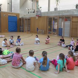Mehrere Mädchen in Sportbekleidung sitzen in einem Kreis auf dem Basketballplatz, einige stehen herum, wahrscheinlich in einer Schulsporthalle.