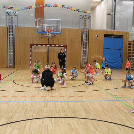 Eine Gruppe von Kindern spielt Fußball auf einem Hallenplatz, beaufsichtigt von einem Erwachsenen. Sie tragen farbenfrohe Trikots und Turnschuhe.