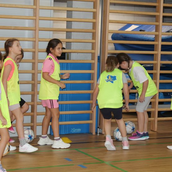 Eine Gruppe junger Mädchen in neonfarbenen Westen übt im Fitnessstudio Fußball. Jedes Mädchen hat einen Fußball und im Hintergrund befinden sich Holzstangen.