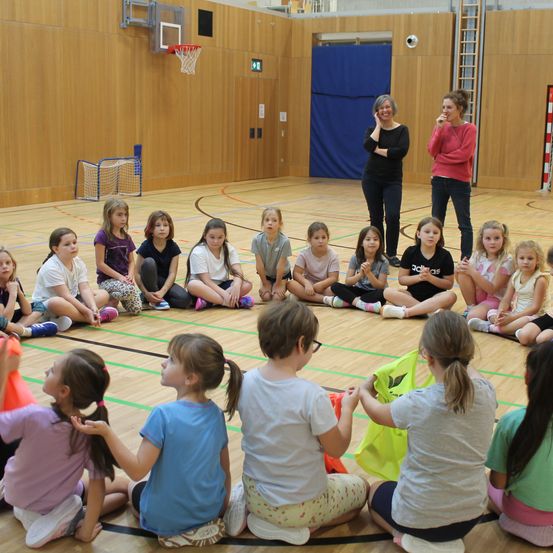 In einer überdachten Turnhalle sitzen eine Gruppe Kinder im Kreis und lauschen aufmerksam zwei Frauen, die in der Mitte stehen. Der Raum ist gut beleuchtet mit Holzwänden und einem Basketballkorb im Hintergrund.