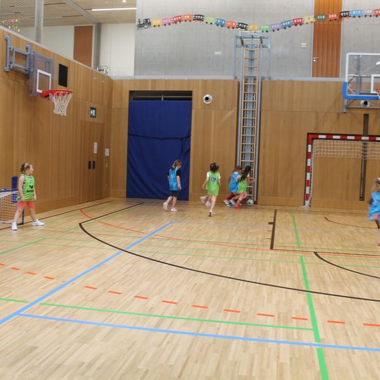 In einer Halle spielen mehrere Kinder Fußball und tragen Teamtrikots. Ein Tor und ein Basketballkorb sind im Hintergrund zu sehen.