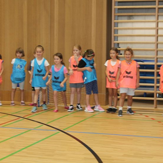 Eine Gruppe junger Mädchen in blauen und orangefarbenen Trikots steht auf einem Basketballplatz, lächelt und schaut nach vorne.