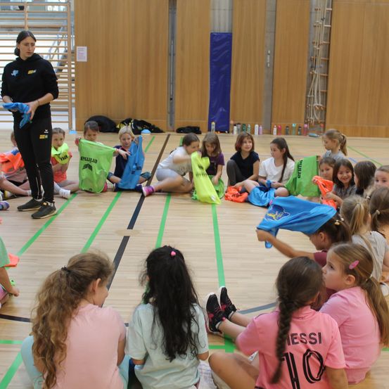 Eine Sporthalle mit Holzboden, in der eine Frau eine Gruppe junger Mädchen beim Training anführt und grüne und blaue Flaggen hält. Die Mädchen sitzen auf dem Boden, einige stehen, alle tragen Nummern auf ihren Trikots. Eine Leiter und Flaschen befinden sich an der Seitenlinie.