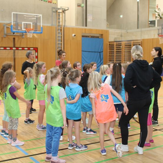 In einer Sporthalle stehen eine Gruppe junger Mädchen in passenden Westen um zwei Frauen herum, von denen eine ein Mikrofon hält. Die Halle verfügt über einen Basketballkorb und ein rotes Netz im Hintergrund.
