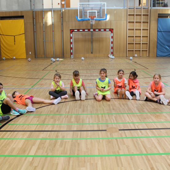 Eine Gruppe junger Kinder in Sportkleidung sitzt und liegt auf einem Basketballplatz, umgeben von Fußballbällen. Dahinter befindet sich ein Basketballkorb und ein Netz, mit einem gelben Vorhang links und einer Holzleiter rechts.