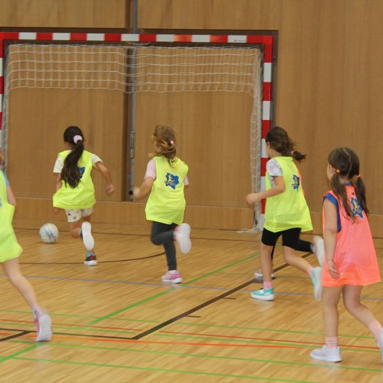 Fünf Mädchen in gelben Westen spielen Hallenfußball. Ein Mädchen läuft dem Ball hinterher, andere folgen. Ein Tor steht dahinter.