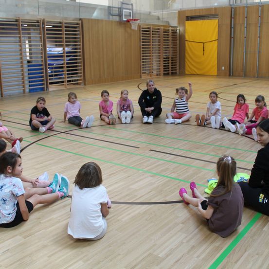 Eine Gruppe von Mädchen sitzt in einem Kreis auf einem Holzboden in einer Turnhalle, mit zwei Frauen auf der Seite. Eine Frau hebt ihre Hand.