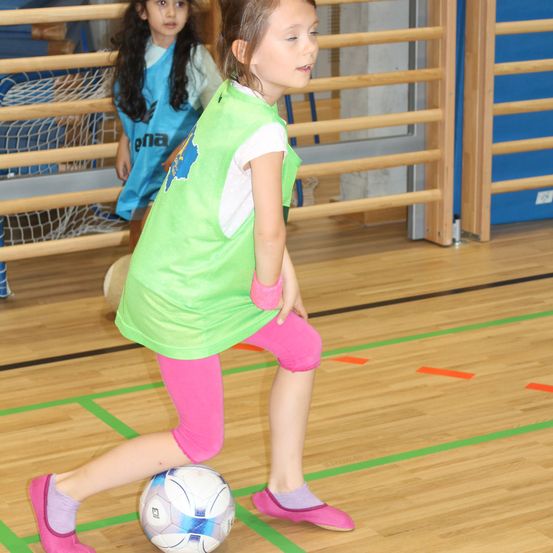 Ein kleines Mädchen in einem grünen Shirt und pinken Hosen spielt Fußball mit einem Ball in einer Sporthalle.