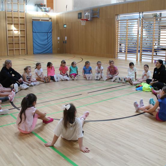 In einer Turnhalle sitzen mehrere Kinder und Erwachsene im Kreis. Eine Frau leitet den Kreis, während die Kinder zusehen.