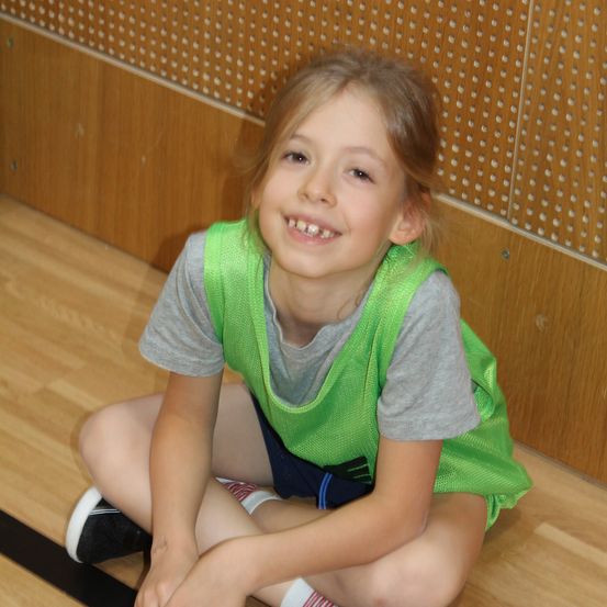 Ein kleines Mädchen mit kurzen Haaren sitzt auf dem Boden und lächelt. Es trägt ein grünes Westchen über einem grauen Shirt.