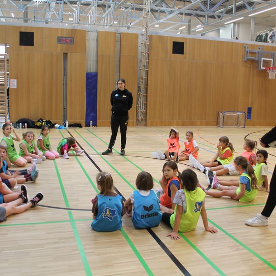 Eine Gruppe junger Mädchen in Sporttrikots sitzt kreisförmig auf dem Hallenboden und hört einer erwachsenen Frau zu, die vor ihnen steht. Der Raum hat Holzwände, ein Basketballkorb und eine Anzeigetafel.