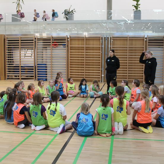 Eine Gruppe junger Mädchen in Sportuniformen sitzt in einem Kreis auf einem Gymnastikboden, mit zwei Frauen in der Nähe. Im Hintergrund befinden sich Holzbarrieren und eine Glaswand, über der Menschen gehen.