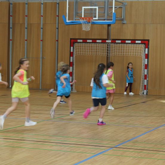 Mehrere Mädchen in farbenfroher Sportbekleidung rennen auf einem Hallenbasketballplatz. Ein Basketballkorb und ein Tor sind im Hintergrund zu sehen.