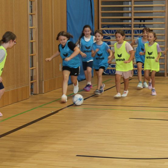 Eine Gruppe von Mädchen in gelben und blauen Trikots spielt Fußball in einer Halle. Sie rennen und jagen einem Ball auf einem Holzboden hinterher.