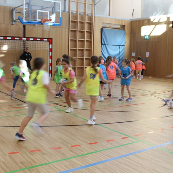 Mehrere junge Mädchen in gelben Westen spielen Basketball auf einem Hallenplatz. Eines der Mädchen hält einen Ball, während die anderen herumlaufen. Eine Erwachsene beobachtet von der Seite.