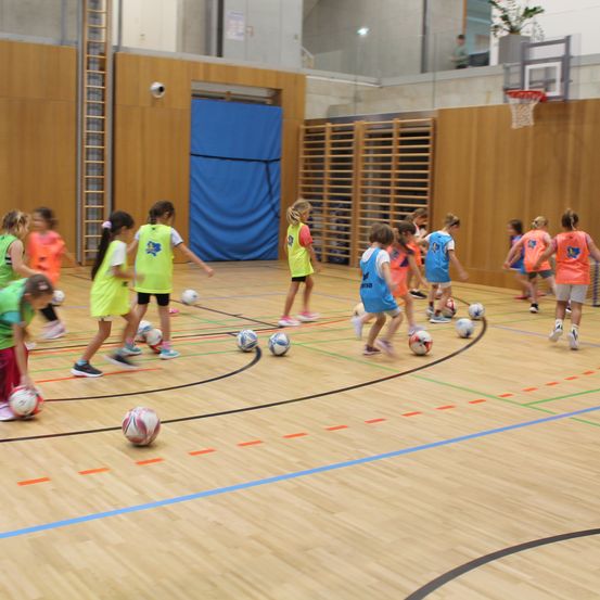 Eine Gruppe von Kindern in farbenfrohen Trikots spielt Fußball auf einem Hallenplatz. Bälle sind auf dem Platz verstreut, und ein Basketballkorb ist im Hintergrund zu sehen.