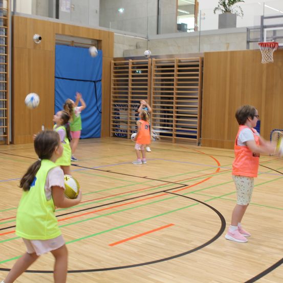 Mehrere Kinder spielen Ball in einer Turnhalle. Sie tragen alle gelbe Westen und Turnschuhe. Der Platz hat einen Basketballkorb auf der rechten Seite.