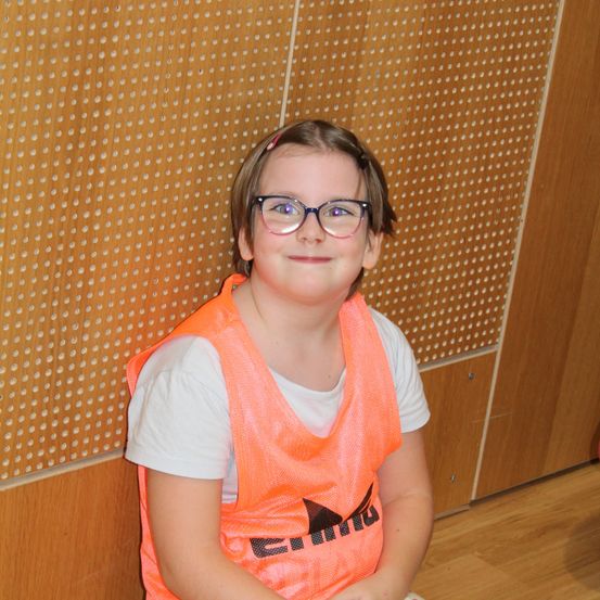 Ein junges Mädchen mit Brille und orangem Weste sitzt lächelnd an einer strukturierter Wand.