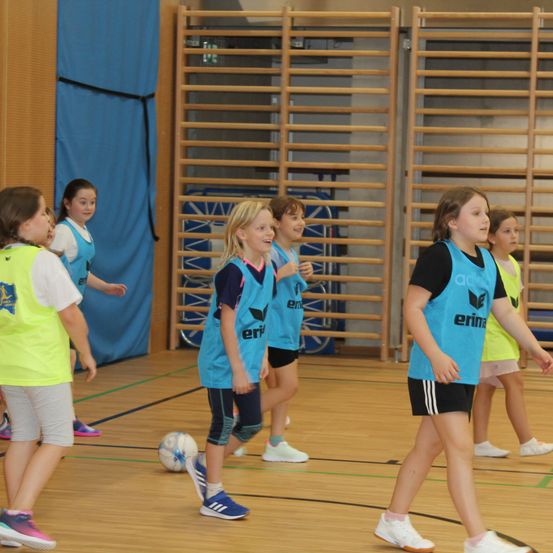 Eine Gruppe junger Mädchen spielt Hallenfußball. Sie tragen passende Trikots und Turnschuhe. Der Platz hat Holzboden und blaue Vorhänge.