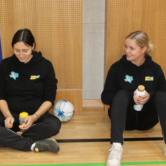 Zwei Frauen in Sportkleidung sitzen lächelnd auf dem Boden, mit einem Fußball und einer Flasche zwischen ihnen.