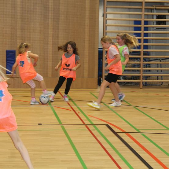 Fünf Mädchen spielen auf einem Hallenplatz Fußball und tragen orangefarbene Westen. Das Mädchen in der Mitte tritt den Ball.