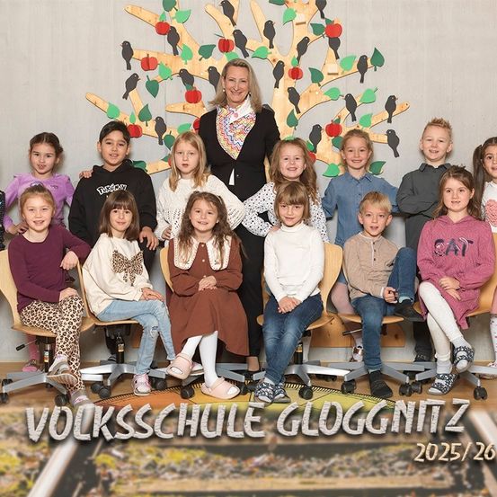 Eine Gruppe von Kindern und ihrer Lehrerin posiert für ein Foto vor einem geschmückten Baum. Der Hintergrund zeigt einen Baum mit Äpfeln und Vögeln. Der Schulname und das Jahr sind unten angegeben.