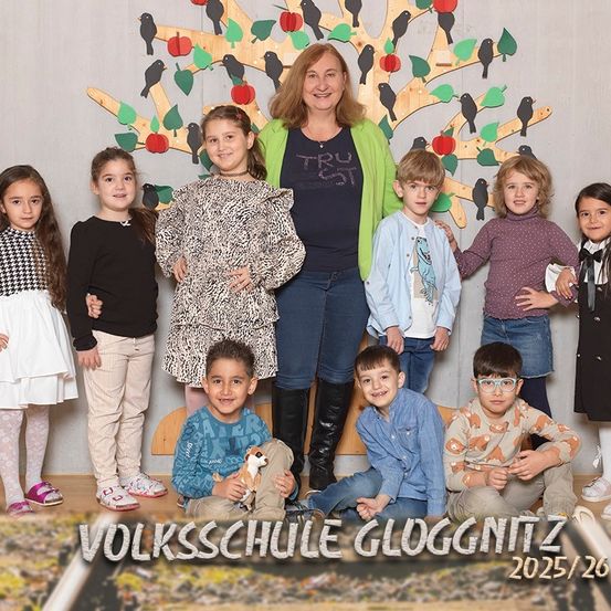 Eine Gruppe von Kindern und einer erwachsenen Frau posiert für ein Foto vor einer dekorierten Baumwand in der Volksschule Gloggnitzer. Unten steht das Jahr 2025/26 geschrieben.
