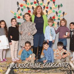 Eine Gruppe von Kindern und einer erwachsenen Frau posiert für ein Foto vor einer dekorierten Baumwand in der Volksschule Gloggnitzer. Unten steht das Jahr 2025/26 geschrieben.