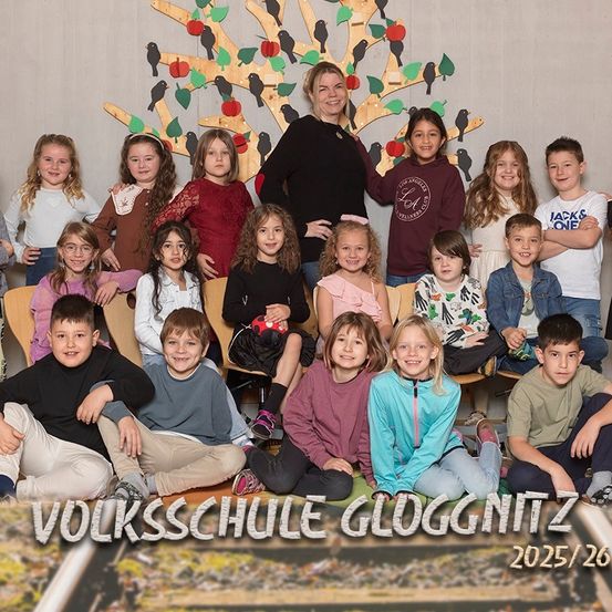 Eine Gruppe von Kindern und einer Frau posiert für ein Foto vor einem geschmückten Baum. Das Bild enthält den Text 'Volksschule Gloggnitz 2025/26'.
