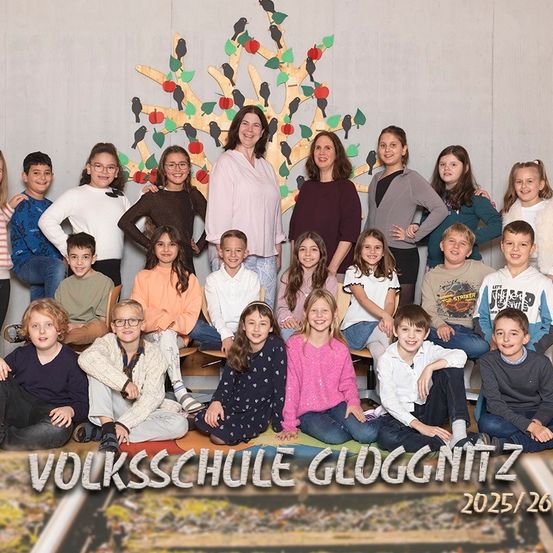 Eine Gruppe von Schülern und zwei Lehrern posiert für ein Foto vor einem geschmückten Baum. Das Bild hat einen Wasserzeichen, der 'VOLKSSCHULE GLOGGNITZ 2025/26' lautet.