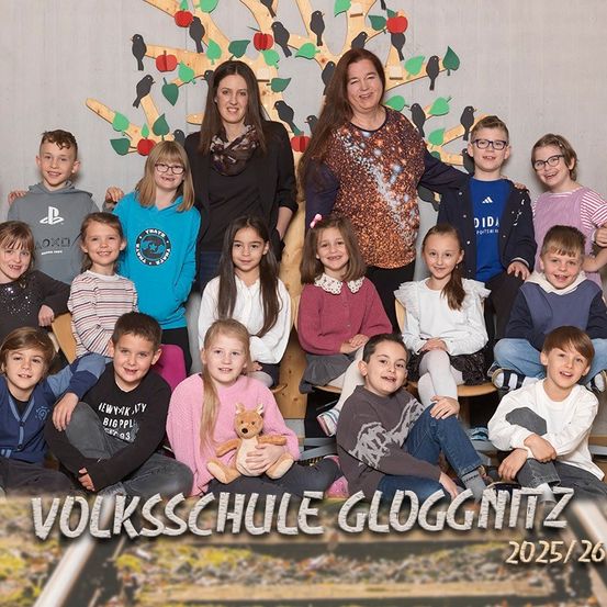 Eine Gruppe von Kindern und zwei erwachsenen Frauen posiert für ein Foto vor einem Baumhintergrund mit Äpfeln und Blättern. Der Text lautet 'Volksschule Gloggnitz 2025/26'.