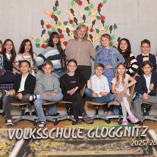 Eine Gruppe von Schülern und einer Lehrerin der Volksschule Gloggnitzer posiert für ein Klassenfoto. Das Foto ist vor einer Baumkulisse mit Äpfeln und Vögeln. Die Bildunterschrift lautet 'Volksschule Gloggnitzer 2025/26'.