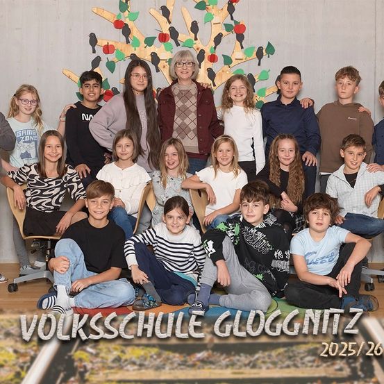 Ein Gruppenfoto von Schülern und einer Lehrerin an der Volksschule Glugginitz für das Jahr 2025/26. Schüler sitzen auf Stühlen und auf dem Boden, während eine Lehrerin in der Mitte mit einem Baumhintergrund steht.